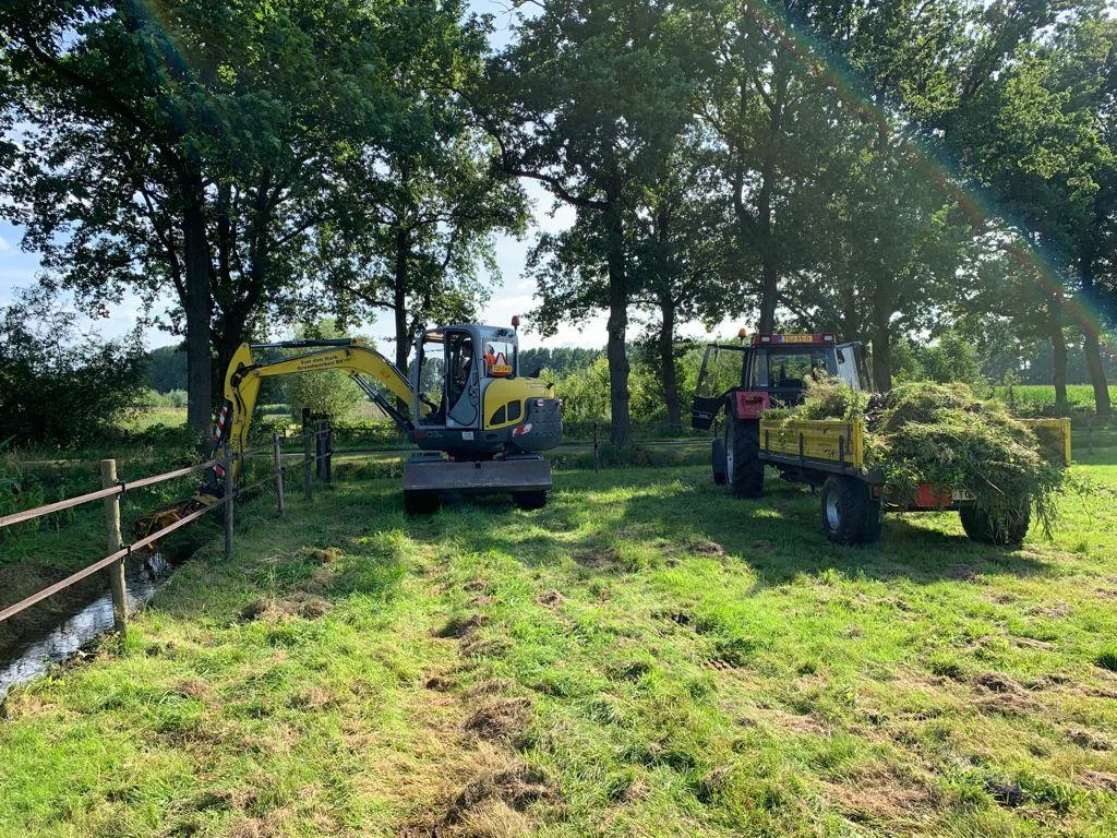 Gele minigraver en tractor met kar op een weiland naast een sloot